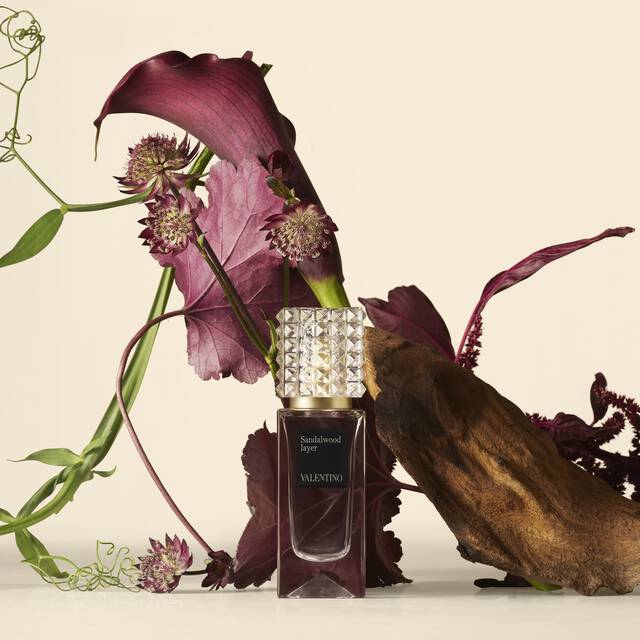 A Valentino perfume bottle placed beside deep red flowers and textured natural elements against a soft, light background.