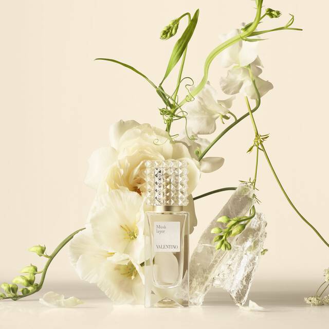 A Valentino perfume bottle arranged with white flowers and translucent crystals on a neutral background.