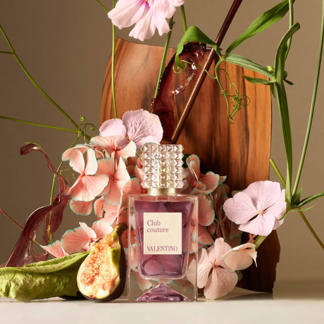 A bottle of Valentino's "Club Couture" perfume is elegantly displayed among vibrant pink flowers and greenery, with a piece of sliced fig nearby and a wooden backdrop.