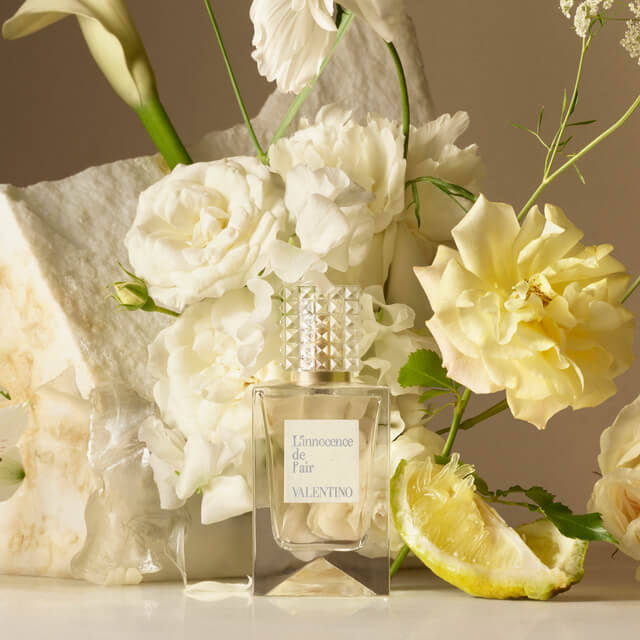 A stylish perfume bottle labeled "L'Innocence de Fair" by Valentino, surrounded by white and yellow flowers and a slice of lemon on a neutral background.