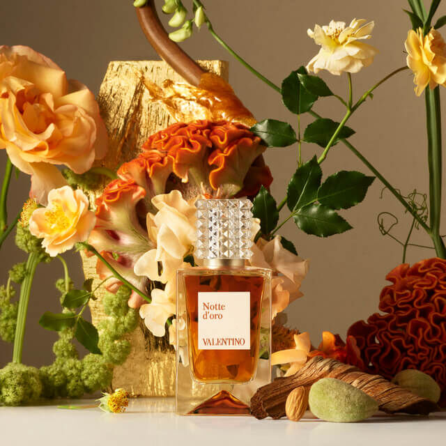 A bottle of Valentino's Notte d'Oro perfume sits elegantly among colorful flowers and foliage, with a golden decorative element in the background.