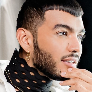 Ein junger Mann mit modischem Haarschnitt und Bart schaut nachdenklich in einen Spiegel, wahrend er einen eleganten Schal tragt.