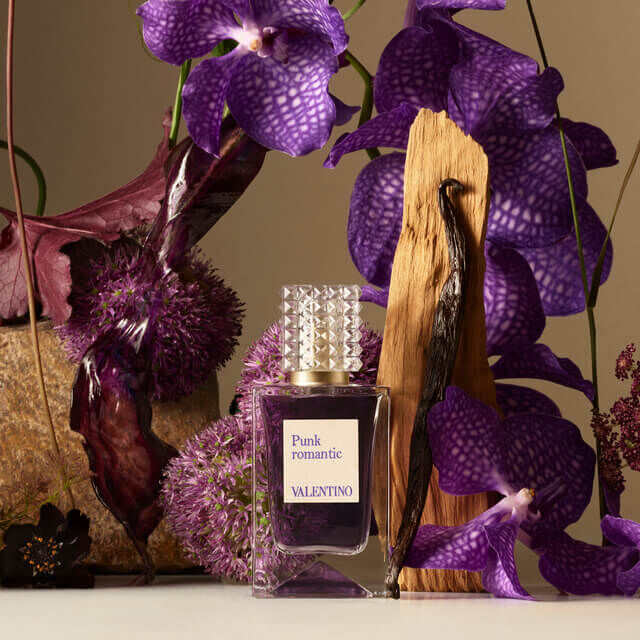 A bottle of Valentino's "Punk Romantic" perfume sits elegantly amidst purple orchids, textured foliage, and vanilla pods, highlighting a luxurious and artistic setting.