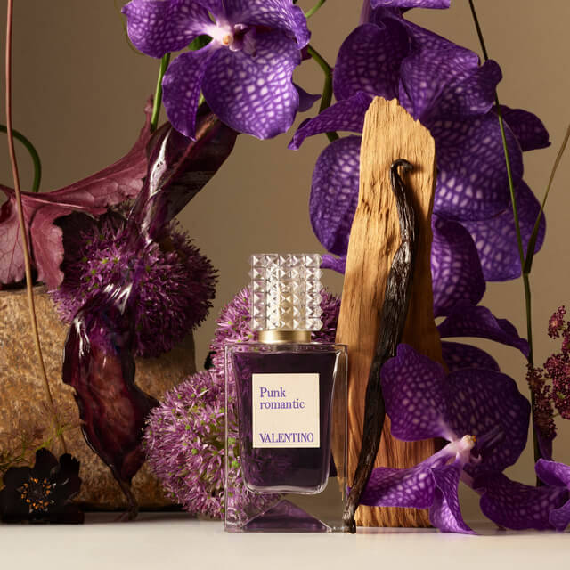 A bottle of Valentino's "Punk Romantic" perfume sits elegantly amidst purple orchids, textured foliage, and vanilla pods, highlighting a luxurious and artistic setting.