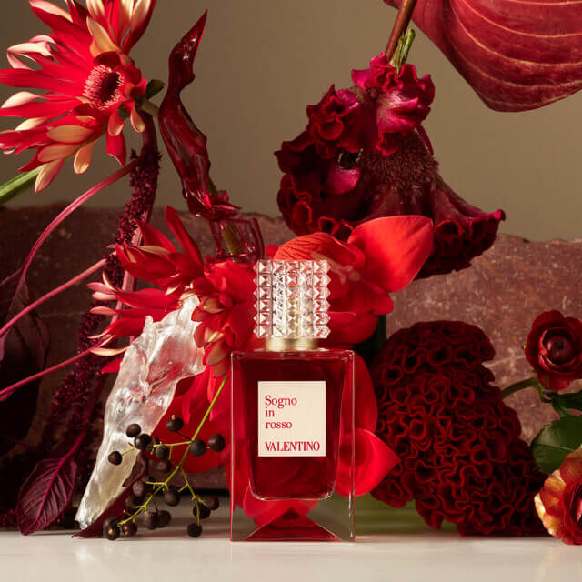 A luxury perfume bottle labeled "Sogno in Rosso" by Valentino, surrounded by vibrant red and burgundy flowers and dramatic foliage.