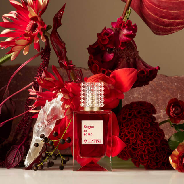 A luxury perfume bottle labeled "Sogno in Rosso" by Valentino, surrounded by vibrant red and burgundy flowers and dramatic foliage.