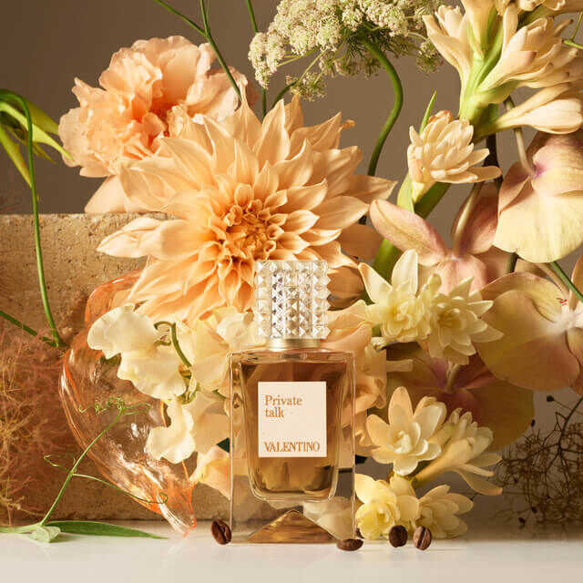 A bottle of Valentino's "Private Talk" perfume surrounded by an arrangement of soft peach and cream flowers.